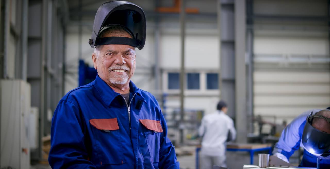 Älterer Mann in blauer Arbeitskleidung und hochgeklapptem Schweißerschutzhelm in einer Industriehalle.