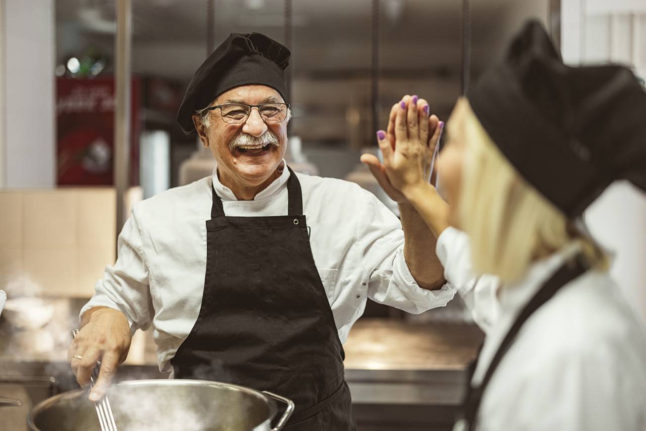 Ein älterer Koch und eine junge Köchin geben sich während der Arbeit in einer Restaurantküche lachend ein High-Five.