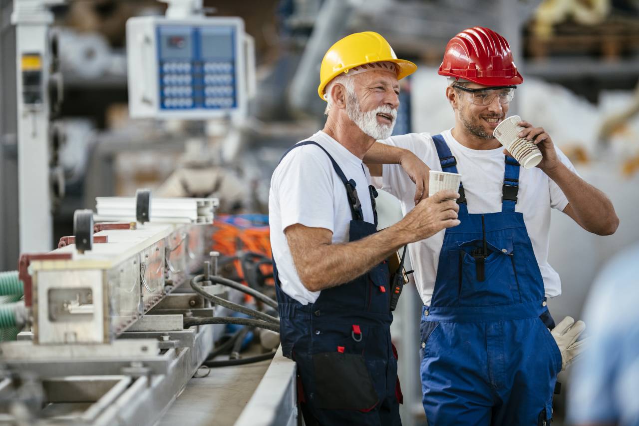 Zwei ältere Handwerker in Arbeitskleidung stehen lachend mit Kaffeetassen in einer Werkstatt zusammen.