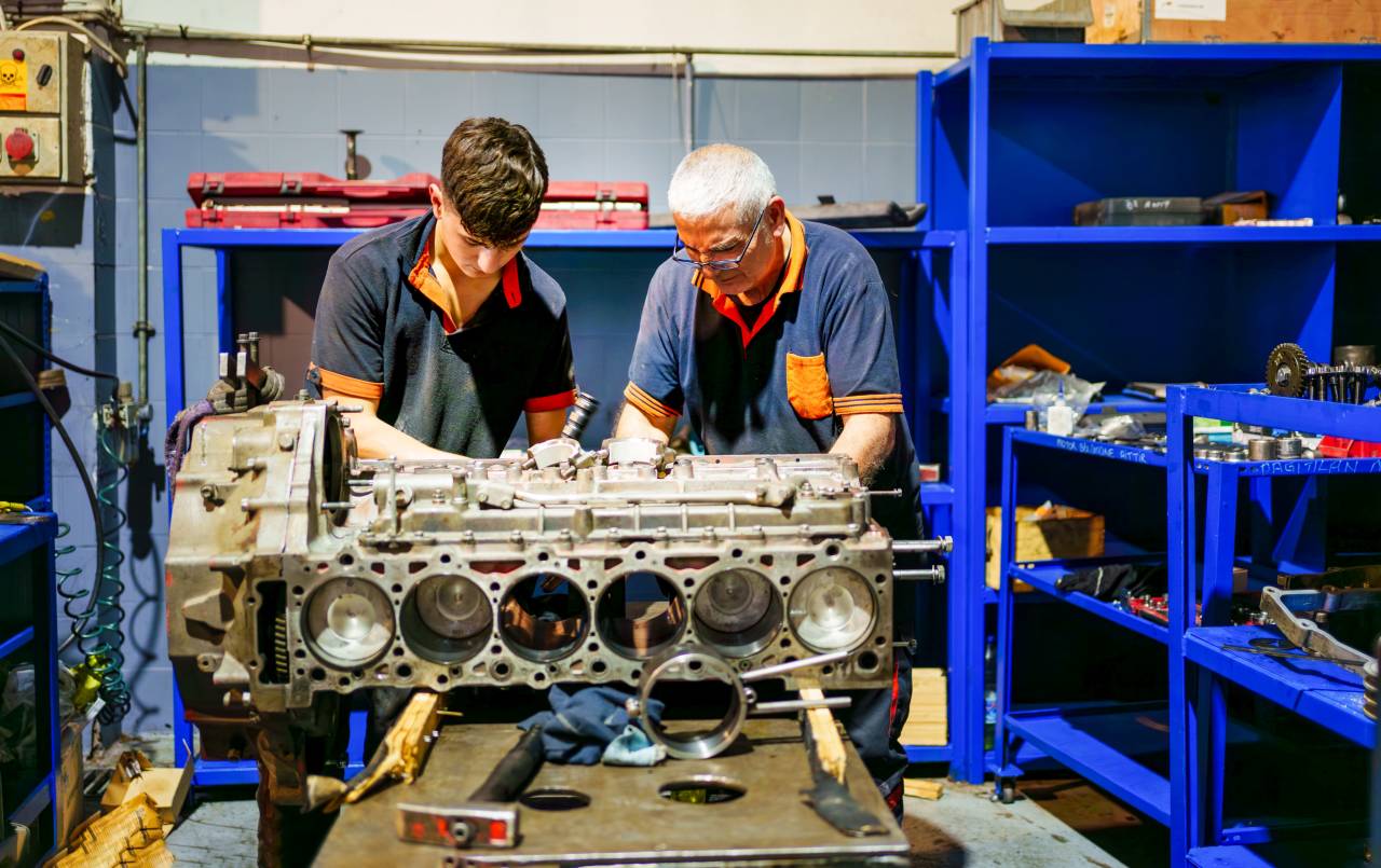 Ein älterer Mitarbeiter arbeitet zusammen mit einem Auszubildenden an einem Motorblock.